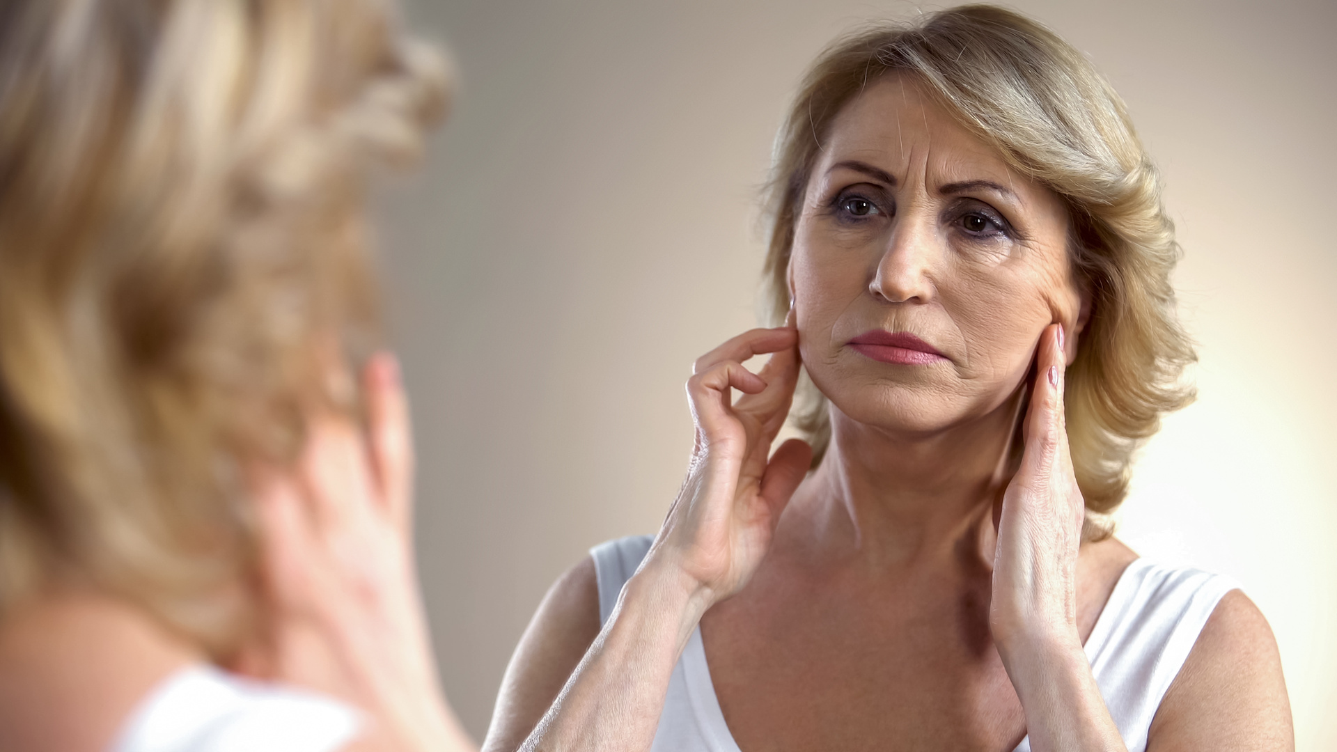 Unhappy aged woman looking in mirror at home, touching face, aging process